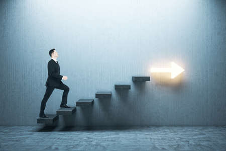 Businessman in suit moving up on stairs with arrow on gray wall. Leadership and career development concept.の写真素材