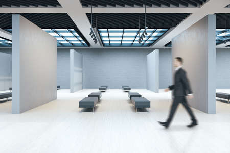 Businessman walking in exhibition room with copyspace on concrete wall and benchs. Gallery and...