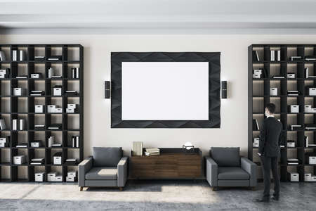 Businessman standing in living room interior with bookshelf and white blank billboard on wall....