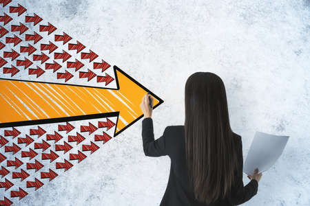 Businesswoman with paper in hand drawing group arrows on concrete wall. Leadership and challenge...