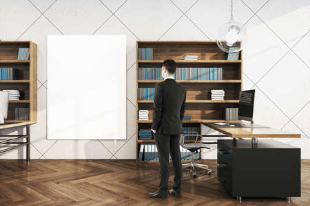 Businessman standing in office interior with computer and looking on blank poster on wall