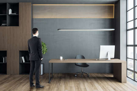 Businessman standing in director's office with computer on wooden table and shelf with documents....