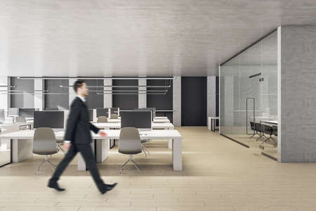 Businessman walking in coworking office interior with computers on table and panoramic city view. Workplace and worker concept.の写真素材
