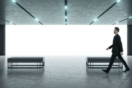 Businessman in black suit walking in a modern exhibition room with blank projection screen and...