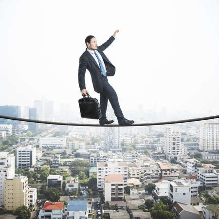 Business risk concept with businessman in black suit with briefcase walking the tightrope over cityの写真素材