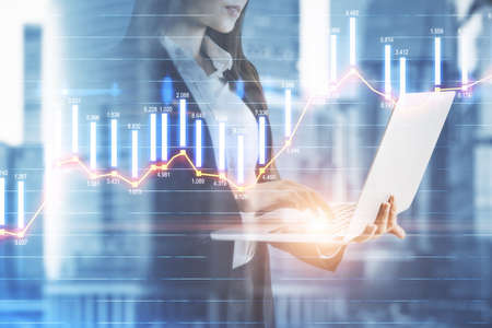 Businesswoman counting the trading bar charts and holding a laptop on the background with city view, market and analysis concept, double exposureの写真素材