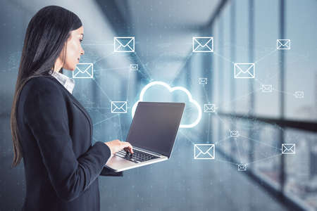 Businesswoman standing and holding a laptop with cloud holo symbol and message icons in the background, business communication and social media conceptの写真素材