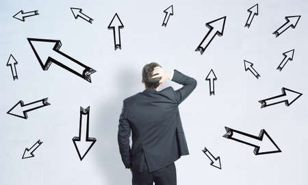 Business decision concept with man in black suit looking at white wall with lots of arrows directed...