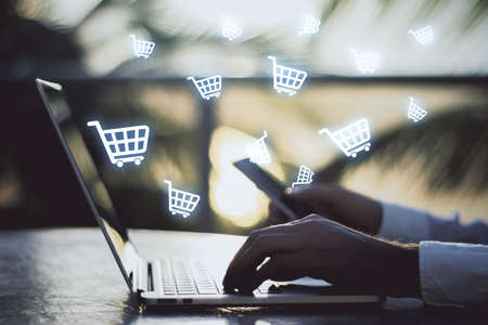 Multiple white shopping carts icons with hands touching the laptop keyboard in the background, side view, online shopping and purchase conceptの写真素材