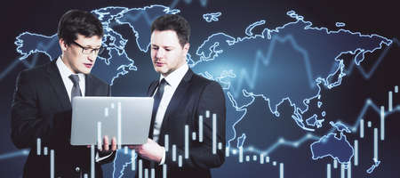 Two businessman with a laptop analyze data with trading charts and a world map in the background,...