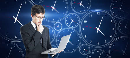 Time management concept. Pensive young european businessman with laptop on creative blue clock...