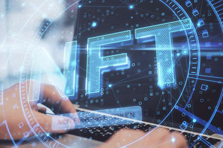 Close up of hands using laptop keyboard with glowing NFT non fungible token concept hologram on...