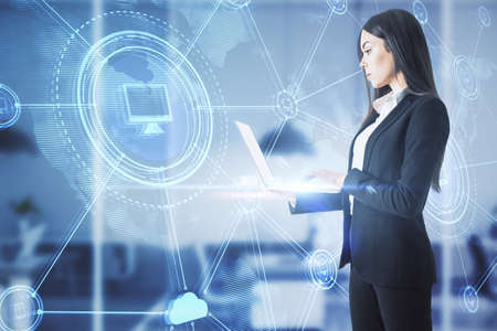 Thoughtful young european businesswoman with laptop standing in blurry office interior with abstract glowing digital business interface. Double exposureの写真素材