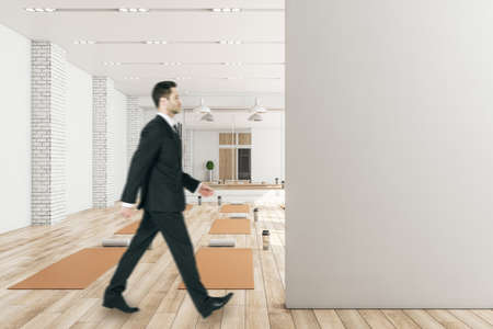 Businessman walking in modern concrete yoga gym interior with equipment, empty mock up place on wall, daylight and wooden flooring. Healthy lifestyle concept. Mock upの写真素材