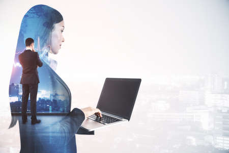 Thoughtful young businesswoman using laptop while standing on abstract white city background with forex chart and mockup place. Brainstorming, technology and market concept. Double exposureの写真素材