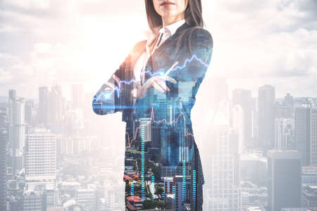 Attractive young european businesswoman with folded arms standing on abstract bright city background with forex chart and mockup place. Finance, trade and success concept. Double exposureの写真素材