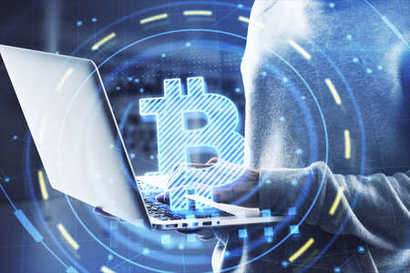 Hacker in hoodie using laptop computer with glowing bitcoin interface in blurry office interior....