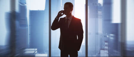 Young businessman talking on the phone in office with panoramic blurry city view. Future, communication, executive and success conceptの写真素材