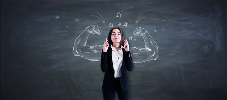 Pretty young european businesswoman with crossed fingers standing on chalkboard wall background with...