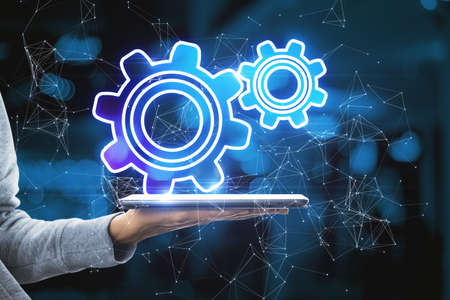 Close up of hand holding tablet with abstract glowing gear wheels on blurry bokeh background with...