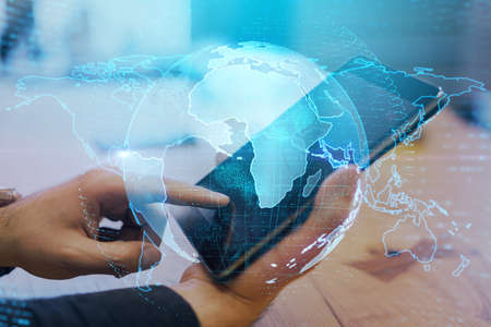 Close up of hands at desktop using tablet with creative glowing globe interface on dark background. Technology, network and online data concept. Double exposureの写真素材