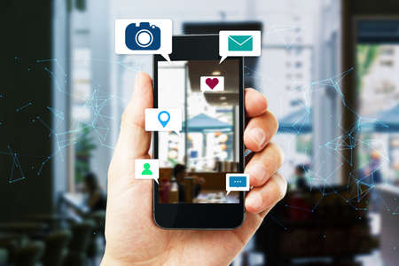 Close up of hand holding smart phone device with abstract polygonal mesh, camera, location pin, email and chat icons on daylight city background. Social media, communication, technology and internet conceptの写真素材