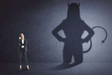 Attractive thoughtful young european businesswoman with devil shadow standing in empty concrete interior. Disguise, demon and career conceptの写真素材