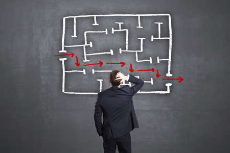 Back view of young man looking at maze and arrow sketch on concrete wall background. Solution,...