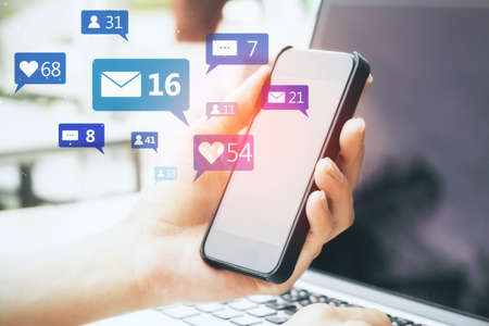 Close up of hand using smartphone and laptop with social media icons on blurry background....