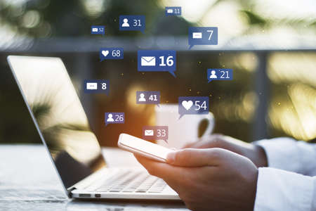 Close up of hand with coffee cup using laptop and smartphone at desktop with social media icons on...