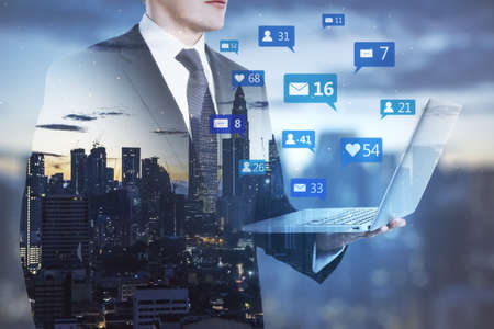 Businessman holding laptop with social media icons on blurry night city background. Communication, network, connections, device and app concept. Double exposureの写真素材
