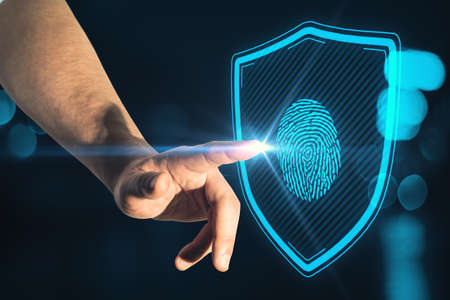 Close up of male hand pointing at creative glowing blue finger print and shield interface on blurry dark bokeh background. Access, technology and identity conceptの写真素材