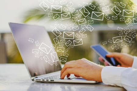 Close up of male hands using smartphone and laptop at desktop with creative drawn email icons on...