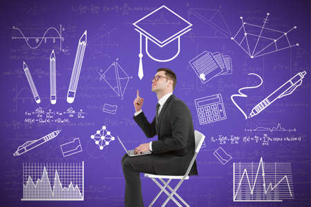 Portrait of attractive young european businessman with laptop sitting and pointing up at creative knowledge and graduation sketch on purple wall background. Education and academic conceptの写真素材