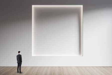 Businessman standing in minimalistic concrete and wooden interior with abstract clean square frame on wall. Mock up. Exhibition, gallery and art conceptの写真素材