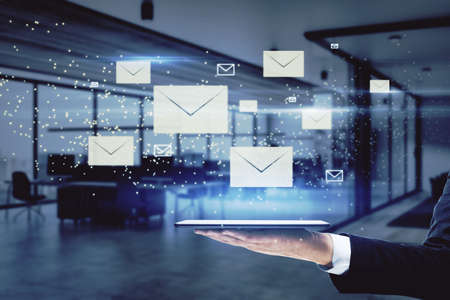 Close up of businessman hand holding pad with creative glowing letters interface on blurry office interior background. E-mail networking and correspondence conceptの写真素材