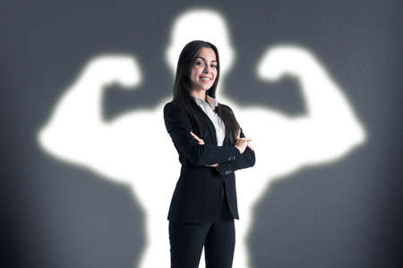 Happy young european businesswoman with creative white muscle arms shadow on concrete wall...