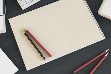 Top view of empty spiral notepad on dark wooden desktop with keyboard and pencils. Drawing,...