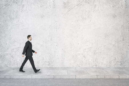 Side view of young businessman walking in concrete interior with mock up place on wall. Success...