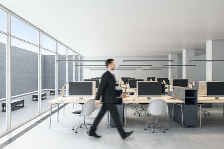 Businessman walking in modern coworking office interior with empty computer monitors on wooden desks, equipment, furniture and panoramic windows with daylightの写真素材