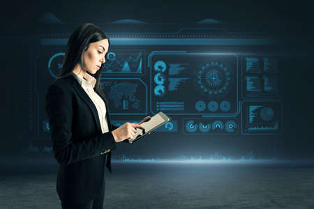 Attractive young european businesswoman using tablet while standing on dark background with glowing digital hud business interface. Future, technology and innovation conceptの写真素材