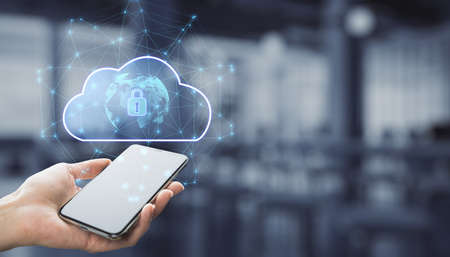 Close up of businessman hand holding smartphone with abstract glowing cloud hologram with globe and safety icons, polygonal mesh on blurry interior background with mock up place. Big data, information, computing and database concept. double exposureの写真素材
