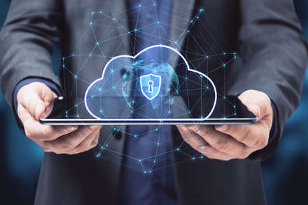 Close up of businessman hand holding tablet with abstract glowing cloud hologram with globe and safety icons, polygonal mesh on blue bokeh interior background. Big data, information, computing and database concept. double exposureの写真素材