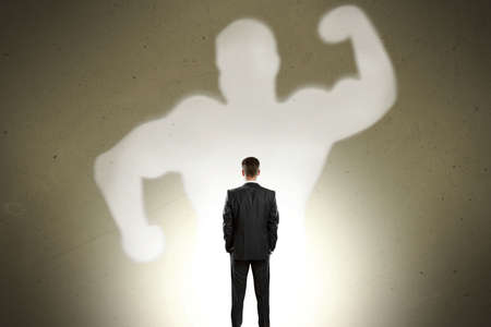 Back view of businessman with strong muscle shadow standing on gray wall background. Confidence and strength conceptの写真素材