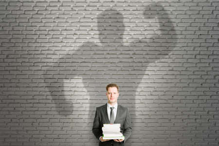 Abstract image of attractive european businessman with books and strong muscle shadow on brick wall background. Confidence and strength conceptの写真素材