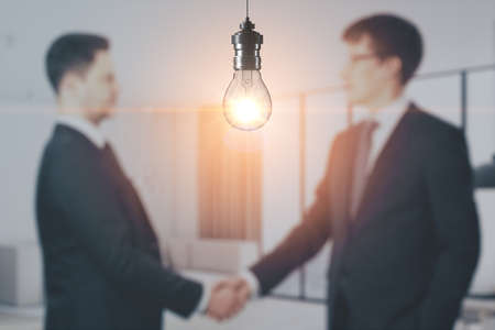 Blurry businesspeople shaking hands together on abstract office interior background with glowing light bulb. Work ideas and innovation concept. Mock up place for your advertisementの写真素材