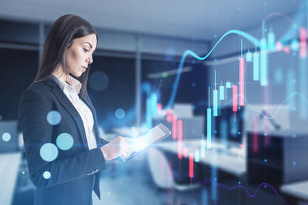 Attractive european businesswoman holding laptop with glowing business chart on blurry office interior background. Trade, finance, market and financial growth concept. double exposureの写真素材