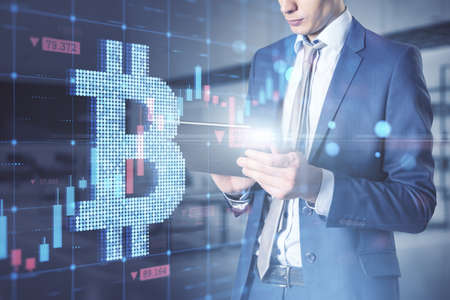 Attractive young caucasian businessman using digital tablet with creative glowing bitcoin grid forex chart hologram on blurry office interior background. Cryptocurrency and growth concept. double exposureの写真素材