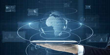 Close up of businessman hand holding tablet with glowing globe and business chart hologram on blue background. World and corporate media concept. double exposureの写真素材
