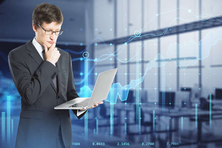 Attractive thoughtful young european businessman with laptop standing in blurry office interior with...